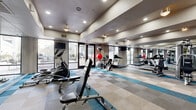 This fitness center is a spacious, single-floor facility thoughtfully designed to accommodate a wide variety of workout styles and fitness goals. The layout is open and well-organized, with distinct zones dedicated to cardio, strength training, and functional fitness. The flooring features a contemporary checkerboard pattern in neutral tones accented with teal stripes, lending the space a modern, energetic aesthetic.

The cardio area is lined with a row of treadmills, elliptical trainers, and stationary recumbent bikes, positioned alongside large windows and glass doors that allow generous natural light to fill the room and offer views to an outdoor area beyond. Full-length mirrors span an entire wall, visually expanding the space and allowing users to monitor their form throughout their workouts.

The strength training section is well-stocked with cable machines, multi-functional weight stations, and a dumbbell rack offering a wide range of free weights. Stability balls and medicine balls are also available, supporting functional and core training routines. Exercise mats are stored neatly for floor-based workouts.

Recessed lighting throughout the ceiling provides consistent, even illumination, while ceiling fans contribute to comfortable air circulation. A vending machine is conveniently located within the space. The tiered, coffered ceiling design adds architectural interest, giving the facility a polished and professional appearance overall.