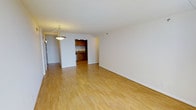 This single-floor space is characterized by its open, airy layout and consistent light hardwood flooring that runs throughout, lending warmth and continuity to the interior. The main living area is generously sized, with smooth white walls and a white ceiling that together enhance the sense of brightness and openness. A pendant light fixture with a gold-toned finish anchors the dining or entryway zone, adding a subtle decorative touch to an otherwise clean, minimalist aesthetic.

Visible through an open passageway, the kitchen features medium-toned wood cabinetry on both upper and lower sections, complemented by what appears to be a dark stone or laminate countertop. The kitchen is compact yet efficiently arranged, with recessed lighting providing focused illumination within the space.

The transition between the main living area and the kitchen is defined by a wide, open archway, maintaining a connected and flowing feel throughout. White baseboards frame the perimeter of each room, providing a crisp finish. Overall, the space presents a neutral, move-in-ready environment with a straightforward and functional design suitable for a variety of uses.