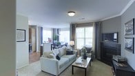 This well-appointed single-floor residence showcases an open-concept layout that effortlessly connects the main living areas, creating a cohesive and functional flow throughout. The living room serves as the centerpiece of the space, featuring plush neutral-toned seating, a glass-top coffee table, and warm carpeting underfoot. Crown molding along the ceiling adds a refined architectural touch, while a ceiling-mounted light fixture provides consistent ambient illumination. Large windows dressed with layered curtains and blinds flood the space with generous natural light, enhancing the bright and airy atmosphere. Adjacent to the living area, the dining zone accommodates a full dining set with dark wood chairs, positioned near an additional set of windows that further amplify the sense of openness. The kitchen is partially visible beyond the dining area, featuring a raised breakfast bar, dark cabinetry, and what appears to be tile flooring, offering a pleasing contrast to the carpeted living and dining spaces. Warm earth tones, taupe walls, and carefully coordinated furnishings contribute to a welcoming and cohesive aesthetic throughout. The overall design balances comfort and style, making it a practical yet visually appealing living environment.