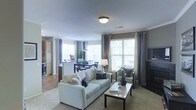 This well-appointed single-floor residence showcases an open-concept layout that effortlessly connects the main living areas, creating a cohesive and functional flow throughout. The living room serves as the centerpiece of the space, featuring plush neutral-toned seating, a glass-top coffee table, and warm carpeting underfoot. Crown molding along the ceiling adds a refined architectural touch, while a ceiling-mounted light fixture provides consistent ambient illumination. Large windows dressed with layered curtains and blinds flood the space with generous natural light, enhancing the bright and airy atmosphere. Adjacent to the living area, the dining zone accommodates a full dining set with dark wood chairs, positioned near an additional set of windows that further amplify the sense of openness. The kitchen is partially visible beyond the dining area, featuring a raised breakfast bar, dark cabinetry, and what appears to be tile flooring, offering a pleasing contrast to the carpeted living and dining spaces. Warm earth tones, taupe walls, and carefully coordinated furnishings contribute to a welcoming and cohesive aesthetic throughout. The overall design balances comfort and style, making it a practical yet visually appealing living environment.