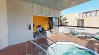 Located in the Encino area of Los Angeles, CA, this rooftop amenity level offers residents a well-rounded set of recreational features under the Southern California sun. The outdoor terrace is finished with classic terra cotta brick pavers and serves as the central hub of the space, connecting all key areas seamlessly.

A built-in spa and hot tub anchors the terrace, providing a relaxing retreat with open-sky exposure. Adjacent to the spa, a dedicated fitness room is enclosed behind sliding glass doors and features stationary bikes, a rowing machine, a multi-station weight bench, and additional exercise equipment, all set against bold yellow accent walls that add energy to the workout space.

The terrace extends to an open-air balcony overlooking a tree-lined neighborhood, complete with wrought iron table-and-chair seating arrangements ideal for outdoor relaxation. Mature eucalyptus and palm trees frame the views, contributing to the lush, California ambiance.

A utility and safety area near the entrance includes posted pool and spa rules, emergency signage, and fire safety equipment, reflecting well-maintained, code-compliant facilities. The space benefits from abundant natural light throughout the day, making it a bright and inviting environment for fitness, leisure, and outdoor enjoyment.