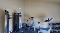This single-floor fitness room is thoughtfully designed to serve as a dedicated workout space. The room features neutral beige walls and dark, durable carpet flooring well-suited for exercise use. It is equipped with a selection of quality fitness machines, including an Inspire cable weight system for strength training, a Precor elliptical trainer for low-impact cardio, and a Precor stationary recumbent bike. The equipment is arranged to make efficient use of the available floor space, allowing for comfortable movement between machines. A ceiling vent ensures adequate air circulation throughout the room, contributing to a comfortable workout environment. The clean, uncluttered layout and neutral color palette create a focused, functional atmosphere suited for regular fitness routines. The space offers everything needed for a well-rounded workout without leaving the building, making it a practical and convenient amenity.