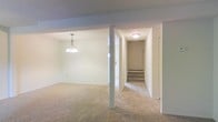 This single-floor space presents a straightforward, functional layout characterized by clean lines and a neutral color palette throughout. The main living area is finished with wall-to-wall beige carpeting, lending a warm and cohesive feel underfoot. A pendant chandelier hangs overhead, providing a focal point and adding a touch of classic character to the space.

A structural column subtly defines the transition between the open living zone and the entry corridor, maintaining an airy flow without fully closing off either area. The entryway hallway is well-lit by a flush-mount ceiling fixture and leads to the front door, accompanied by a short staircase that connects to the building's common areas or exterior access point.

The textured ceilings are consistent throughout, complementing the white and light gray wall tones that keep the space feeling bright and open. The layout is efficient, with clear sightlines from the main living area to the entry corridor. Overall, the space offers a clean, move-in-ready environment with a simple, understated aesthetic that provides a neutral canvas for personalization.