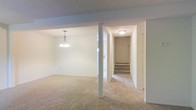 This single-floor space presents a straightforward, functional layout characterized by clean lines and a neutral color palette throughout. The main living area is finished with wall-to-wall beige carpeting, lending a warm and cohesive feel underfoot. A pendant chandelier hangs overhead, providing a focal point and adding a touch of classic character to the space.

A structural column subtly defines the transition between the open living zone and the entry corridor, maintaining an airy flow without fully closing off either area. The entryway hallway is well-lit by a flush-mount ceiling fixture and leads to the front door, accompanied by a short staircase that connects to the building's common areas or exterior access point.

The textured ceilings are consistent throughout, complementing the white and light gray wall tones that keep the space feeling bright and open. The layout is efficient, with clear sightlines from the main living area to the entry corridor. Overall, the space offers a clean, move-in-ready environment with a simple, understated aesthetic that provides a neutral canvas for personalization.