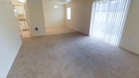 This single-story space offers an open and airy layout designed for comfortable everyday living. The main living area features neutral-toned wall-to-wall carpeting that extends throughout, creating a cohesive and understated aesthetic. A set of vertical blinds flanks a large sliding glass door or window, allowing generous natural light to fill the room while offering a connection to the outdoors. The open floor plan flows naturally into an adjoining area distinguished by light-toned hard flooring, providing a subtle visual separation between living zones without compromising the sense of openness. A simple pendant light fixture hangs in the adjoining space, adding a clean, functional accent overhead. The walls are finished in a soft, neutral tone that complements the flooring and contributes to the overall bright and welcoming atmosphere. A small window on the far wall introduces additional natural light into the secondary area. The layout is straightforward and versatile, lending itself well to a variety of furniture arrangements and personal styling choices. Overall, the space presents a clean, move-in-ready environment with a calm, neutral palette throughout.