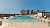 This generously sized outdoor pool area is designed to serve as a central amenity and social hub. The pool itself is a standout feature, showcasing crystal-clear blue water with built-in water jets and a tanning ledge complete with submerged chaise loungers, allowing residents to relax partially in the water. A series of stepping stone pavers bisect a shallow wading section, adding both visual interest and functional access across the pool.

The surrounding sundeck is finished in warm-toned concrete pavers and is lined with white chaise lounges on multiple sides, providing ample space for sunbathing. A substantial wood pergola anchors one end of the deck, offering shaded outdoor seating beneath its open-beam lattice roof. Beneath the pergola sits a well-appointed outdoor kitchen area with a built-in grill and stone-clad counters, complemented by wicker dining chairs and round tables ideal for al fresco gatherings.

Adjacent to the pool, a modern farmhouse-style clubhouse building provides an additional covered lounge area. The entire space is enclosed by a tall cedar privacy fence and black iron fencing, with manicured green lawn areas adding softness to the hardscape surroundings.