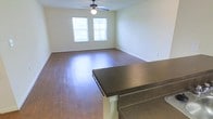 This single-floor space offers a clean, functional layout centered around an open living area. The room features rich hardwood flooring that extends throughout, reflecting natural light that enters through two windows dressed with white horizontal blinds. The windows provide a bright, airy atmosphere while maintaining privacy. A ceiling fan with an integrated light fixture is mounted centrally overhead, ensuring year-round comfort and adequate illumination. The walls are finished in a neutral, light tone that complements the warm wood flooring and contributes to the overall sense of openness. A dark-topped breakfast bar with decorative white support columns serves as a natural divider between the living area and what appears to be an adjacent kitchen space. The bar's dark countertop contrasts attractively with the lighter surroundings, adding a visual anchor to the open-plan layout. White baseboards run along the perimeter of the room, providing a crisp finish. The overall ambiance is practical and inviting, with a straightforward design that offers a versatile, move-in-ready living environment suited to a range of furnishing styles.