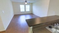 This single-floor space offers a clean, functional layout centered around an open living area. The room features rich hardwood flooring that extends throughout, reflecting natural light that enters through two windows dressed with white horizontal blinds. The windows provide a bright, airy atmosphere while maintaining privacy. A ceiling fan with an integrated light fixture is mounted centrally overhead, ensuring year-round comfort and adequate illumination. The walls are finished in a neutral, light tone that complements the warm wood flooring and contributes to the overall sense of openness. A dark-topped breakfast bar with decorative white support columns serves as a natural divider between the living area and what appears to be an adjacent kitchen space. The bar's dark countertop contrasts attractively with the lighter surroundings, adding a visual anchor to the open-plan layout. White baseboards run along the perimeter of the room, providing a crisp finish. The overall ambiance is practical and inviting, with a straightforward design that offers a versatile, move-in-ready living environment suited to a range of furnishing styles.