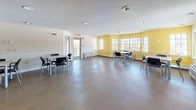 This community room is a generously sized, single-floor gathering space designed to serve a variety of group activities and social functions. The main area features durable hard-surface flooring and a textured ceiling with recessed lighting, creating a bright and functional environment. Multiple tables paired with modern black chairs are arranged throughout the space, providing flexible seating configurations suitable for meetings, events, or casual gatherings.

One of the room's standout features is a curved accent wall finished in a warm yellow tone, complemented by a row of large multi-pane windows that invite ample natural light and outdoor views. A secondary door provides direct exterior access, enhancing convenience for events. The open layout allows for easy reconfiguration to suit different needs.

A dedicated kitchenette area supports the space with dark espresso cabinetry, a stainless steel refrigerator, a wall oven, and a sink — offering practical amenities for catered gatherings or community use. White wainscoting along the lower walls adds a clean, classic architectural detail throughout. The overall ambiance is welcoming and versatile, balancing functional design with comfortable, community-oriented features.