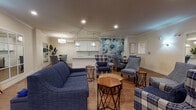 Located at 30 Main in Londonderry, NH, this single-floor community space is designed with both comfort and functionality in mind. The open-concept layout features warm hardwood flooring throughout, crown molding, and recessed lighting that create a welcoming and polished ambiance.

The central lounge area is generously sized and furnished with plush navy blue sofas, plaid accent chairs, and geometric side tables, all arranged to encourage conversation and relaxation. A striking watercolor floral mural in shades of blue serves as a bold focal point, adding an artistic touch to the space. Adjacent to the lounge is a dedicated media area with a wall-mounted flat-screen television, pendant lighting, and comfortable seating.

The kitchenette features sleek white cabinetry, a light glass tile backsplash, stainless steel appliances, and a large white countertop island illuminated by modern pendant lights — ideal for casual gatherings and refreshments. A separate dining nook with a wood table and upholstered chairs sits beside the mural wall, offering an intimate setting.

A dedicated activity room with large framed mirrors, wall sconces, and an open floor plan provides a versatile space for fitness or wellness programs. The accessible restroom is finished with decorative patterned floor tile, dark subway tile wainscoting, textured wall tile, and stainless steel grab bars, combining style with practicality.