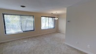 This single-floor space offers a clean and functional layout designed for comfortable everyday living. The main living area is generously proportioned, featuring soft wall-to-wall carpeting that adds warmth underfoot and a neutral, inviting atmosphere throughout. Walls are finished in a light beige tone, creating a bright and airy feel that complements the natural light streaming in through multiple large windows fitted with horizontal blinds. These windows frame views of lush, mature trees outside, seamlessly connecting the interior with the surrounding natural environment. A stylish pendant light fixture hangs as a focal point within the space, adding a touch of elegance to the otherwise understated interior. Crisp white baseboards provide a clean contrast against the carpeted floors and neutral walls, contributing to the space's polished, well-maintained appearance. A wall-mounted thermostat is also visible, indicating climate control provisions for year-round comfort. The open layout allows for versatile furniture arrangement, making the space adaptable to a variety of uses. Overall, the space exudes a calm, neutral ambiance with practical features and ample natural light, making it a welcoming and functional environment.