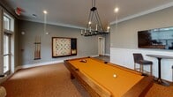 Located at 557 Roanoke Rd in Daleville, Virginia, this single-floor recreation and game room is thoughtfully designed to offer a well-rounded leisure experience. The space is anchored by a solid wood pool table with a bold amber felt surface, suspended beneath an elegant wrought-iron candelabra-style chandelier that serves as the room's centerpiece. Recessed lighting complements the chandelier, ensuring the area is well-lit throughout. A full-size shuffleboard table lines one wall, adding another dimension of entertainment to the room. A large wall-mounted flat-screen TV is positioned above a wainscoted accent wall, paired with a high-top bistro table and cushioned bar stools — ideal for casual viewing or conversation. A wall-mounted oversized Scrabble board adds a playful, decorative touch. Large windows dressed with colorful patterned drapes and horizontal blinds flood the space with natural light and offer views of the surrounding outdoors. The warm cork-toned carpet grounds the room with a cohesive, inviting feel, while the cool gray walls and crisp white wainscoting lend a clean, modern aesthetic. A wide corridor with tile flooring connects the game room to additional areas of the facility, reinforcing the functional and social nature of this community-oriented space.