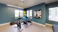 This fitness studio is thoughtfully designed to support a variety of exercise routines in a clean, modern setting. The space is defined by bold teal accent walls that contrast with crisp white ceilings and baseboards, creating a visually stimulating environment conducive to an active workout. A smooth, durable light-toned floor runs throughout, providing a practical surface suitable for both equipment use and floor exercises.

Natural light flows in through large black-framed windows that frame lush greenery and palm trees outside, bringing an airy, refreshing quality to the room. A ceiling fan assists with ventilation and comfort during intense sessions. Floor-to-ceiling mirrors along one wall visually expand the space and allow users to monitor their form, while a wall-mounted flat-screen TV adds an entertainment element.

The studio is equipped with multiple stationary spin bikes arranged for group cycling sessions, along with an assortment of colorful exercise balls and medicine balls stored on a dedicated rack. A wall-mounted yoga mat holder keeps the space organized. A smart control panel is mounted on the wall for convenient climate or audio-visual management. Black-framed glass double doors connect the studio to adjacent areas, enhancing the open, cohesive feel of the overall facility.