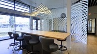 This single-floor space showcases a distinctly contemporary design aesthetic, blending functional workspaces with striking architectural details. At its center sits a large, natural wood communal table surrounded by modern adjustable bar-height seating in charcoal gray, creating an inviting collaborative hub. Overhead, a dramatic sculptural light fixture composed of angled linear LED tubes commands attention, while a contrasting dark spherical pendant adds visual depth nearby.

A standout feature is the intricate white lattice partition wall, whose undulating wave-like pattern serves as both a functional divider and an artistic installation, creating a dynamic interplay of light and shadow throughout the space. Light hardwood flooring runs underfoot, complementing the warm tones of the wood table and contributing to an open, airy feel.

Large floor-to-ceiling windows along the street-facing wall flood the interior with abundant natural light and offer clear views to the exterior, seamlessly connecting the inside with the outdoors. The overall ambiance is polished and professional, yet approachable — well-suited for collaborative work, meetings, or client-facing interactions.