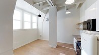 This single-floor space combines industrial character with a clean, modern aesthetic. The open layout is anchored by exposed ceiling beams and structural columns, which lend an architectural interest while preserving the airy, loft-like atmosphere. The crisp white walls and ceilings amplify the natural light that pours in through multi-pane windows, creating a bright and welcoming environment throughout the day. Light hardwood flooring runs continuously across the space, adding warmth and a sense of cohesion to the interior. Pendant lighting — including a distinctive black dome fixture and a classic white shade — hangs from the beamed ceiling, providing both functional illumination and stylish focal points. A partial wall with built-in shelving offers subtle spatial division without compromising the open feel. A glimpse of a kitchen area to one side reveals white cabinetry and a countertop, seamlessly integrated into the overall design. The combination of industrial structural elements with a neutral, light-filled palette creates a versatile and inviting space well-suited to a variety of uses.
