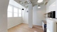 This single-floor space combines industrial character with a clean, modern aesthetic. The open layout is anchored by exposed ceiling beams and structural columns, which lend an architectural interest while preserving the airy, loft-like atmosphere. The crisp white walls and ceilings amplify the natural light that pours in through multi-pane windows, creating a bright and welcoming environment throughout the day. Light hardwood flooring runs continuously across the space, adding warmth and a sense of cohesion to the interior. Pendant lighting — including a distinctive black dome fixture and a classic white shade — hangs from the beamed ceiling, providing both functional illumination and stylish focal points. A partial wall with built-in shelving offers subtle spatial division without compromising the open feel. A glimpse of a kitchen area to one side reveals white cabinetry and a countertop, seamlessly integrated into the overall design. The combination of industrial structural elements with a neutral, light-filled palette creates a versatile and inviting space well-suited to a variety of uses.