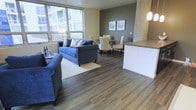 This single-floor space features an open-concept layout that seamlessly connects the kitchen, dining, and living areas, creating a cohesive and functional environment. Rich, dark hardwood flooring extends throughout, providing a warm and grounding contrast to the lighter wall tones. The living area is anchored by a large area rug and benefits from generous natural light streaming through floor-to-ceiling windows along one wall, offering views of the surrounding exterior. A charcoal accent wall with framed artwork adds depth and visual interest to the dining zone, where a round table with upholstered chairs creates an intimate setting. Pendant lighting with frosted globe shades hangs above the kitchen peninsula, which features dark cabinetry and a neutral countertop surface. The peninsula serves as both a functional workspace and a natural visual divider between the kitchen and the living space without disrupting the open flow. A textured stone or tile column detail adds an architectural element between the living and kitchen zones. Overall, the space presents a contemporary aesthetic with a balanced mix of neutral tones, natural materials, and modern lighting.