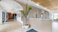 This thoughtfully designed lobby level serves as the communal heart of the building, offering residents a range of functional and social spaces under one roof. At the entrance, large black-framed glass doors open into a bright entryway with large-format tile flooring and a warm wood herringbone-patterned ceiling detail, setting an elevated tone from the moment of arrival. A sleek reception desk in light oak anchors the front lobby, illuminated by a brass chandelier and complemented by vertical wood slat accent walls that lend a refined, coastal aesthetic throughout.

Adjacent to the entry, a generously sized lounge area invites residents to gather, featuring mid-century modern seating arrangements in soft neutral tones, a striking brass multi-arm chandelier, and floor-to-ceiling windows that frame outdoor views and flood the space with natural light. A built-in bench with teal upholstery adds a pop of color against warm wood paneling.

The mailroom area showcases herringbone hardwood flooring, a teal accent wall housing an extensive bank of resident mailboxes, package lockers, and elevator access. A dedicated co-working or community room offers a communal high-top table with bold green seating, a large wall-mounted display screen, and direct outdoor views. Throughout, curated artwork, recessed lighting, and cohesive finishes create a polished and welcoming environment.