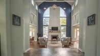 Located at 638 River Bend Ct in Newport News, Virginia, this beautifully appointed single-floor clubhouse delivers an impressive blend of architectural drama and coastal charm. The centerpiece of the space is a grand great room with soaring double-height ceilings, bold navy blue accent walls, and striking white structural truss detailing that draws the eye upward. A floor-to-ceiling arched window frames lush outdoor views, while a classic fireplace with a white shiplap surround and wood mantel anchors the room. Warm hardwood-style flooring runs throughout, complementing a sophisticated seating arrangement of gray sofas and wingback chairs arranged on a neutral area rug. Adjacent to the main lounge, a sunroom-style dining area is bathed in natural light from large windows on multiple walls, offering views of an outdoor deck and surrounding trees. Round tables with wicker chairs create a casual, welcoming atmosphere. A stylishly designed kitchenette features rich navy cabinetry with gold hardware, white quartz countertops, textured tile backsplash, and coastal-inspired artwork, providing a fully functional refreshment area. Throughout the space, recessed lighting, exposed trusses, and an abundance of windows ensure the interiors remain bright, airy, and connected to the natural surroundings.