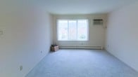 This single-floor space presents a clean and straightforward layout designed for comfortable living. The main living area is finished with soft, light-gray wall-to-wall carpeting that adds warmth underfoot and contributes to the room's understated, neutral aesthetic. Walls are painted in a crisp white, enhancing the sense of brightness and openness throughout.

A prominent picture window serves as the focal point of the main room, allowing generous amounts of natural light to fill the interior while offering a view of the surrounding brick buildings and greenery outside. Beneath the window runs a baseboard heating unit, providing an efficient and evenly distributed heat source during cooler months. A wall-mounted air conditioning unit positioned in the upper corner of the room ensures year-round climate control.

The overall design is minimal and functional, with smooth ceilings and clean wall lines that give the space a tidy, well-maintained character. The neutral color palette throughout makes the space highly adaptable for a variety of furnishing styles. The combination of natural light, practical climate systems, and carpeted flooring creates a livable and inviting environment.