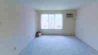 This single-floor space presents a clean and straightforward layout designed for comfortable living. The main living area is finished with soft, light-gray wall-to-wall carpeting that adds warmth underfoot and contributes to the room's understated, neutral aesthetic. Walls are painted in a crisp white, enhancing the sense of brightness and openness throughout.

A prominent picture window serves as the focal point of the main room, allowing generous amounts of natural light to fill the interior while offering a view of the surrounding brick buildings and greenery outside. Beneath the window runs a baseboard heating unit, providing an efficient and evenly distributed heat source during cooler months. A wall-mounted air conditioning unit positioned in the upper corner of the room ensures year-round climate control.

The overall design is minimal and functional, with smooth ceilings and clean wall lines that give the space a tidy, well-maintained character. The neutral color palette throughout makes the space highly adaptable for a variety of furnishing styles. The combination of natural light, practical climate systems, and carpeted flooring creates a livable and inviting environment.