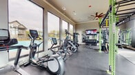 The fitness center at The Preserve in Athens, Georgia, is a spacious, single-floor facility thoughtfully designed to support a comprehensive workout experience. The room is bathed in natural light, courtesy of expansive floor-to-ceiling windows that line the walls and offer views of the surrounding outdoor pool area and wooded landscape, creating an energizing and open atmosphere.

The facility is organized into distinct training zones. A dedicated cardio area runs along the windowed perimeter and features a well-stocked row of treadmills, elliptical trainers, stationary bikes, and cycling bikes equipped with individual tablet screens. A functional training zone anchors the center of the room with bold green power racks, a suspended rig for bodyweight and resistance training, and battle ropes, all set atop durable rubber mat flooring. A strength and free weights section provides a full dumbbell rack, cable machines, and additional resistance equipment.

Multiple wall-mounted flat-screen televisions are positioned throughout the space for entertainment during workouts. Large decorative ceiling fans with distinctive blade designs add to the room's visual character while maintaining airflow. Recessed lighting complements the natural light throughout. Smooth, durable flooring transitions between workout zones, and a designated stretching and functional movement area features padded sport turf matting. Double glass entry doors provide an inviting entrance to this well-appointed amenity.