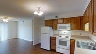 This single-floor space showcases a practical and well-organized layout, with the kitchen serving as a central highlight. The kitchen is outfitted with warm-toned wood cabinetry that provides generous storage both above and below the countertops. White appliances, including a top-freezer refrigerator, an electric coil range, and an over-the-range microwave, offer a clean, cohesive look. Light-colored countertops complement the cabinetry, creating a balanced contrast throughout the space. A textured ceiling adds subtle visual interest overhead, while a multi-bulb chandelier-style light fixture provides warm, focused illumination. Dark hardwood-style flooring runs throughout, grounding the space with a rich, durable surface. HVAC vents are integrated into both the ceiling and lower wall areas, ensuring efficient climate control. An open passage from the kitchen leads directly into an adjoining living area, enhancing the sense of flow and connectivity within the unit. The living area features clean white walls, continuing the bright and neutral palette established in the kitchen. Overall, the space presents a functional and straightforward design suited for everyday living.