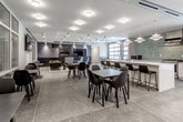 Located along I-29 N in North Kansas City, Missouri, this single-floor amenity space is thoughtfully designed to blend functionality with contemporary style. The open-concept layout encompasses a variety of distinct zones that flow seamlessly into one another, creating a versatile environment for gathering, dining, and relaxing.

The kitchen is a standout feature, outfitted with a large white island with an integrated sink, full-size stainless steel appliances, a microwave, and a bold geometric-patterned tile backsplash. Pendant lighting adds warmth to the space, while ample counter space supports both casual and more involved culinary use.

Adjacent dining areas feature sleek tables paired with modern black chairs set atop large-format stone-look tile flooring. Wall-mounted flat-screen televisions are positioned throughout, providing functional entertainment options for occupants. Glass partition walls bordering the dining zone offer visual transparency while defining separate areas.

A dedicated lounge area provides a more relaxed atmosphere, anchored by a dramatic dark wood accent wall with an inset fireplace. A mustard-toned sofa, lounge chairs, and accent tables create an inviting setting. Large glass garage-style doors flood this portion of the space with natural light and offer views to the exterior. A separate area with a warm wood ceiling and polished concrete flooring adds further design variety, completing this well-appointed and multifunctional space.