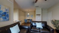This single-floor space offers a well-organized, open-concept layout that seamlessly connects the main living areas. The living room serves as the heart of the space, anchored by a dark leather sectional sofa adorned with blue geometric accent pillows, creating a comfortable and inviting atmosphere. Decorative beams add subtle architectural character to the ceiling above.

Adjacent to the living area, a compact dining nook accommodates a small table paired with upholstered blue chairs beneath a classic pendant light, making it an ideal spot for casual meals. Blue floral artwork on the walls ties the color palette together and adds a lively, decorative touch throughout the space.

A wall-mounted coat hook rack near the entry provides practical organization near what appears to be the main entrance. The kitchen is partially visible, featuring a countertop bar that opens toward the living and dining areas, enhancing the sense of connectivity and flow.

Throughout the space, warm neutral wall tones are complemented by wood-look flooring, contributing to a cohesive and welcoming ambiance. Ceiling vents and recessed fixtures ensure functional climate control. The overall design balances everyday practicality with thoughtful decorative details, resulting in a comfortable and livable single-floor space.
