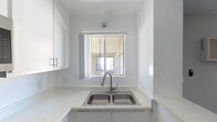 This single-floor space showcases a crisp, minimalist aesthetic throughout. The kitchen is thoughtfully designed with bright white cabinetry featuring sleek bar-pull hardware, offering generous storage above and below the countertops. The counters are finished in a speckled white stone or quartz-style surface, providing a clean and durable workspace. A stainless steel double-basin sink is centrally positioned, equipped with a modern pull-down faucet. A sliding window positioned directly above the sink allows natural light to filter in, brightening the space and offering an outward view. The all-white walls and cabinetry create a cohesive, airy atmosphere, while the recessed corner layout of the kitchen maximizes functional workspace. Overall, the space conveys a clean, neutral palette that provides a versatile and well-lit environment.