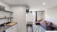 This efficiently designed studio in Lexington, Kentucky, offers a thoughtfully organized single-floor layout that maximizes every square foot. The main living area combines sleeping and lounging in one open space, featuring a full bed, a sectional sofa, and a patterned area rug anchoring the room. A wall-mounted TV and a dedicated dining nook with a compact table and chairs add to the unit's functional versatility. Natural light enters through a window dressed with dark curtains, while recessed lighting and a wall-mounted mini-split system ensure year-round comfort. The kitchenette is neatly tucked along one wall, equipped with open shelving, dark countertops, a sink with a matte black faucet, a two-burner cooktop, a mini refrigerator, and a microwave — providing all essential cooking conveniences. The entry door features a sleek, modern design that complements the unit's neutral gray and white color palette throughout. The bathroom is finished with clean, white subway-tiled walls, a stand-up shower with matte black fixtures, and a minimalist floating sink. Terrazzo-style flooring ties the space together with a contemporary touch, making this studio a practical and well-appointed living space.