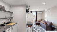 This efficiently designed studio in Lexington, Kentucky, offers a thoughtfully organized single-floor layout that maximizes every square foot. The main living area combines sleeping and lounging in one open space, featuring a full bed, a sectional sofa, and a patterned area rug anchoring the room. A wall-mounted TV and a dedicated dining nook with a compact table and chairs add to the unit's functional versatility. Natural light enters through a window dressed with dark curtains, while recessed lighting and a wall-mounted mini-split system ensure year-round comfort. The kitchenette is neatly tucked along one wall, equipped with open shelving, dark countertops, a sink with a matte black faucet, a two-burner cooktop, a mini refrigerator, and a microwave — providing all essential cooking conveniences. The entry door features a sleek, modern design that complements the unit's neutral gray and white color palette throughout. The bathroom is finished with clean, white subway-tiled walls, a stand-up shower with matte black fixtures, and a minimalist floating sink. Terrazzo-style flooring ties the space together with a contemporary touch, making this studio a practical and well-appointed living space.