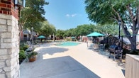 This outdoor amenity area in Lewisville centers around a generously sized, freeform swimming pool with clear turquoise water, complemented by an adjoining spa. The expansive concrete pool deck provides ample room for lounging, furnished with multiple chaise lounge chairs and round tables topped with teal market umbrellas for shaded seating. Mature trees surround the perimeter, offering natural shade and a lush, green backdrop against the open Texas sky. A black iron security fence encloses the pool area, ensuring privacy and safety. Nearby, a covered gazebo-style structure provides additional shelter and a gathering spot for residents. The surrounding buildings feature brick and neutral-toned exteriors with pitched rooflines, contributing to the established character of the community. Potted plants and decorative landscaping add color and visual interest throughout the space. The overall ambiance is open, bright, and inviting, well-suited for outdoor recreation and relaxation in a warm Texas climate.