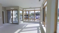 This single-floor space is defined by its striking wall of wood-framed windows that wrap around the main living area, flooding the interior with natural light and offering open views of the surrounding urban neighborhood. The clean, white interior palette — carried across the walls, ceiling, and light-toned flooring — creates a bright and airy atmosphere that feels open and welcoming.

The main area is generously proportioned, with an open layout that maximizes the impact of the panoramic windows. Roller shades are installed on the windows, allowing for flexible light and privacy control while preserving the contemporary aesthetic. A subtle step elevation separates a secondary alcove space from the main floor, adding architectural dimension without disrupting the overall open feel.

Natural light cascades across the smooth floors throughout the day, creating dynamic patterns that shift with the sun. Additional windows along the side wall further enhance ventilation and light. The overall design emphasizes simplicity, openness, and connectivity to the outdoors, making it a functional and visually engaging space suited for a variety of uses.
