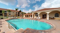 This single-story community space centers around a generously sized outdoor swimming pool with vivid turquoise water, framed by a decorative paver pool deck. The pool reaches a depth of six feet and is equipped with entry railings, making it both accessible and inviting. A row of white lounge chairs lines the deck, offering ample space for sunbathing and relaxation. The poolside clubhouse structure is designed in a Mediterranean-influenced architectural style, featuring warm stucco exteriors in a soft yellow tone, arched colonnades, and a terra cotta hip roof. Decorative lantern-style light fixtures accent the archways, adding a classic touch to the façade. French doors provide access to interior amenities within the clubhouse. The surrounding landscape includes mature palm trees that enhance the tropical atmosphere, while the open sky above allows for abundant natural light throughout the day. Multi-story residential buildings are visible in the background, providing context for the broader community setting. Overall, the space exudes a relaxed, resort-like ambiance well-suited for outdoor leisure and social gatherings.
