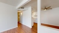 This single-floor, 1-bedroom, 1-bathroom residence offers a practical and thoughtfully organized layout. Warm wood-look flooring runs throughout the main living areas, complemented by crisp white walls and textured ceilings that give the space a clean, bright aesthetic. The open living room is generously proportioned for its footprint, featuring a ceiling fan with light fixture and a large window dressed with vertical blinds that allows natural light to pour in. The galley-style kitchen is efficiently designed with white cabinetry lining both walls, offering ample upper and lower storage. White appliances including a range and refrigerator are included, and a flush-mount ceiling light keeps the space well-lit. A hallway connects the main living areas to the private bedroom and bathroom. The bedroom is a comfortable size, finished with plush carpeting, a window with natural light, and a dedicated closet with built-in shelving and hanging rods. The full bathroom features wood-look flooring, a white vanity with storage, a toilet, and a bathtub with a sliding glass shower enclosure. A connecting hallway with warm accent lighting completes the layout, adding a welcoming sense of transition throughout the home.