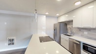 This single-floor space showcases a clean, modern aesthetic with an open-concept design that seamlessly connects the living and kitchen areas. The kitchen is a standout feature, equipped with high-gloss white cabinetry, a large white quartz island with a built-in induction cooktop, and a full suite of stainless steel appliances including a French door refrigerator, dishwasher, and built-in microwave. A marble-look tile backsplash adds a refined touch, complemented by recessed lighting and a sleek pendant light over the island. The open living area is generously proportioned, featuring warm-toned luxury vinyl plank flooring, neutral gray walls, and a ceiling fan. Large windows invite ample natural light throughout the space. The bedrooms continue the consistent flooring and neutral color palette, each offering good natural light through well-placed windows. The bathroom is finished with floor-to-ceiling large-format white subway tiles surrounding a soaking tub and shower combination, accented by chrome fixtures and a small window for ventilation. Overall, the space presents a cohesive, contemporary renovation with durable finishes and a bright, airy atmosphere.