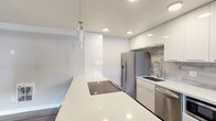 This single-floor space showcases a clean, modern aesthetic with an open-concept design that seamlessly connects the living and kitchen areas. The kitchen is a standout feature, equipped with high-gloss white cabinetry, a large white quartz island with a built-in induction cooktop, and a full suite of stainless steel appliances including a French door refrigerator, dishwasher, and built-in microwave. A marble-look tile backsplash adds a refined touch, complemented by recessed lighting and a sleek pendant light over the island. The open living area is generously proportioned, featuring warm-toned luxury vinyl plank flooring, neutral gray walls, and a ceiling fan. Large windows invite ample natural light throughout the space. The bedrooms continue the consistent flooring and neutral color palette, each offering good natural light through well-placed windows. The bathroom is finished with floor-to-ceiling large-format white subway tiles surrounding a soaking tub and shower combination, accented by chrome fixtures and a small window for ventilation. Overall, the space presents a cohesive, contemporary renovation with durable finishes and a bright, airy atmosphere.