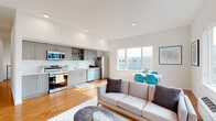 This single-floor unit showcases a clean, contemporary design with an open-concept living, dining, and kitchen layout that maximizes functionality and flow. The kitchen is outfitted with sleek gray cabinetry, white quartz countertops, stainless steel appliances including a range, microwave, dishwasher, and refrigerator, and an open display shelf that adds visual interest. Warm hardwood-style flooring runs throughout the main living areas, complementing the bright white walls and recessed lighting overhead.

The living area is generously proportioned, filled with natural light from large windows offering views of the surrounding neighborhood. A dedicated dining space sits adjacent to the living room, creating a cohesive entertaining area. The bedrooms feature plush carpeting, tall ceilings, and multiple windows that invite abundant daylight. One bedroom includes a walk-in closet with wire shelving systems offering ample storage.

The bathroom maintains the unit's modern aesthetic with gray cabinetry, a white vanity, a large mirror, and hardwood-style flooring. Throughout, the design emphasizes a neutral palette, clean lines, and practical finishes, resulting in a bright, well-organized, and inviting living space.