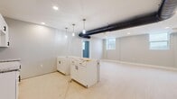 This newly constructed single-floor unit offers a well-designed layout spread across three distinct areas. The open-concept kitchen zone serves as a central hub, featuring white shaker-style cabinetry, a large center island, wall-mounted upper cabinets, and granite countertops. A series of pendant lights hang above the island, complemented by recessed ceiling lighting throughout. A prominent exposed HVAC duct runs across the ceiling, adding an industrial-modern accent to the space. Light-toned flooring flows throughout the unit, contributing to a bright and cohesive aesthetic.

The spacious main living area benefits from multiple windows that allow natural light to filter in, and is equipped with a ceiling fan. Neutral-toned walls and clean white baseboards give the space a fresh, move-in-ready feel. A set of white double doors with black hardware provides access to additional storage or utility areas.

The bedroom is generously sized and features two double-door closets with matching black hardware, offering ample storage. The bathroom is finished with a granite-topped white vanity, an undermount sink, a matte black faucet, and tile flooring, rounding out the unit with a polished, contemporary touch.