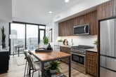 This single-floor residence showcases a thoughtfully designed contemporary layout, beginning with a clean, well-lit entry hallway finished in warm wood-toned flooring and crisp white walls. The corridor leads to an open-concept living and dining area anchored by dramatic floor-to-ceiling windows that frame sweeping urban skyline views, flooding the space with natural light.

The living area features a comfortable seating arrangement oriented toward the city panorama, complemented by modern furnishings, curated artwork, and a dark media console with glass-front cabinetry. The kitchen is equipped with rich dark wood cabinetry, stainless steel appliances including a gas range, built-in microwave, and multi-door refrigerator, set against a classic white tile backsplash with a butcher block island surface.

The bedroom offers a calm, neutral palette with soft carpeting, warm ambient lighting, and framed artwork, creating a restful retreat. The bathroom is finished with large-format tile flooring, a shower-tub combination with tiled surround, ample storage, and notably includes a stacked in-unit washer and dryer. Throughout the residence, the design balances modern aesthetics with functional, livable spaces.