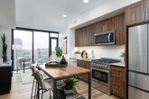 This single-floor residence showcases a thoughtfully designed contemporary layout, beginning with a clean, well-lit entry hallway finished in warm wood-toned flooring and crisp white walls. The corridor leads to an open-concept living and dining area anchored by dramatic floor-to-ceiling windows that frame sweeping urban skyline views, flooding the space with natural light.

The living area features a comfortable seating arrangement oriented toward the city panorama, complemented by modern furnishings, curated artwork, and a dark media console with glass-front cabinetry. The kitchen is equipped with rich dark wood cabinetry, stainless steel appliances including a gas range, built-in microwave, and multi-door refrigerator, set against a classic white tile backsplash with a butcher block island surface.

The bedroom offers a calm, neutral palette with soft carpeting, warm ambient lighting, and framed artwork, creating a restful retreat. The bathroom is finished with large-format tile flooring, a shower-tub combination with tiled surround, ample storage, and notably includes a stacked in-unit washer and dryer. Throughout the residence, the design balances modern aesthetics with functional, livable spaces.