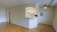 This single-floor space showcases an open-concept design that seamlessly integrates the living area with a pass-through kitchen. The kitchen is framed by a half-wall peninsula with a rounded corner, creating a natural division between the cooking and living zones while maintaining an open, airy feel. White cabinetry lines the upper portion of the kitchen, providing ample storage, and is complemented by a countertop sink, microwave, and refrigerator. A skylight above the kitchen floods the cooking area with natural light, brightening the space considerably. The living area features classic parquet wood flooring in a warm, neutral tone, adding texture and character underfoot. A ceiling fan is mounted overhead, offering both practical air circulation and a functional lighting element. The walls are finished in a clean, neutral palette throughout, contributing to the bright and open ambiance. An electrical panel is mounted on the wall near the kitchen, and white baseboards frame the perimeter of the space, providing a crisp, finished look. Overall, the layout is practical and well-organized, making efficient use of the available space.
