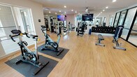 Located at 1160 Montgomery Dr in Allen, TX, this modern fitness center offers a comprehensive workout facility designed to accommodate a wide range of exercise needs. The single-floor space features light hardwood-style flooring throughout, recessed lighting, and a clean, bright aesthetic accentuated by white walls and structural columns that define distinct workout zones.

The cardio section is equipped with multiple treadmills, elliptical machines, stationary cycling bikes, and stair climbers, all positioned near large windows that allow ample natural light to fill the space. Wall-mounted flat-screen televisions are strategically placed throughout the facility, providing entertainment during workouts.

The strength training area includes a variety of cable and selectorized machines, a leg press, and dedicated benches for free weight exercises. A full dumbbell rack lines one wall, complemented by floor-to-ceiling mirrors that enhance the sense of space and allow users to monitor their form.

The facility also features an open stretching and functional training area with ample floor space. Black-framed glass partition doors add a sleek, contemporary touch, separating areas while maintaining visual openness. Ceiling fans and a well-maintained HVAC system ensure comfortable conditions throughout. The overall ambiance is modern, clean, and professional, making this fitness center a well-rounded facility for all fitness levels.