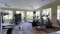 This fitness center is designed to support a well-rounded workout experience within a bright and welcoming environment. The space is finished with warm beige walls, crown molding, and a combination of hardwood-style and carpet flooring that adds texture and visual interest underfoot. A ceiling fan anchors the center of the room, providing airflow and comfort during workouts.

Natural light pours in through multiple large windows along the walls, framing views of lush tropical foliage and palm trees outside, creating an energizing and open atmosphere. A wall-mounted flat-screen television is positioned for easy viewing during cardio sessions.

The layout is thoughtfully arranged to accommodate a diverse selection of equipment. Cardio machines — including treadmills, elliptical trainers, and a stair climber — are positioned to take advantage of the natural light and outdoor views. A dedicated strength and resistance training area features a multi-station cable machine and a weight bench, supporting a full range of strength exercises. Recessed lighting supplements the natural light, ensuring the space remains well-lit at all hours. Overall, this fitness center offers a comfortable, functional, and inviting environment for daily exercise.