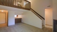 This two-story space offers a practical and open layout that makes efficient use of its vertical design. The main floor features a carpeted living area that flows openly toward a kitchen section, which is equipped with dark wood cabinetry, a countertop, and a sink beneath a window that allows natural light to filter in. The transition between the living and kitchen areas is defined by a raised countertop bar, adding both functionality and visual separation without closing off the space.

A wooden staircase with a warm brown finish rises along one wall, leading up to the second level. The underside of the staircase is cleanly integrated into the wall, keeping the main floor uncluttered. The upper level presents a loft-style overlook, enclosed by a sturdy wooden baluster railing that matches the staircase in tone and material, creating a cohesive design element throughout.

The walls throughout are finished in a neutral off-white, contributing to a bright and airy atmosphere. The open vertical space between the two floors enhances the sense of volume and scale, making the interior feel more expansive than a single-level layout might suggest. Overall, the space combines a straightforward, functional design with warm wood accents that provide a grounded, welcoming character.