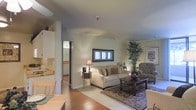 This single-floor residence offers a thoughtfully designed open-concept layout that promotes a sense of flow and connectivity throughout the living spaces. The living room serves as the heart of the home, featuring warm hardwood flooring, neutral-toned walls, and ample space for comfortable seating arrangements. A framed window provides natural light, brightening the room and enhancing its welcoming ambiance. The living area transitions smoothly into the dining space, which is anchored by a wooden dining table, reinforcing the home's warm and cohesive aesthetic. The kitchen, visible from the main living area, is outfitted with white cabinetry and counter space, offering a clean and functional workspace. A structural column adds an architectural element that subtly defines the boundary between the kitchen and living zones without interrupting the open feel. Throughout the space, careful attention to detail is evident in the decorative touches, including a patterned area rug, an ornamental indoor tree, and tasteful accent lighting from a classic table lamp. The overall ambiance is calm, comfortable, and inviting, making this a versatile space well-suited for both everyday living and casual entertaining.