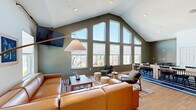 Located at 111 Cochran Rd in Clemson, South Carolina, this single-floor space offers a well-designed layout ideal for gathering and collaboration. The standout feature is the living room, which boasts soaring vaulted ceilings and a dramatic floor-to-ceiling window wall that floods the interior with natural light and frames views of the surrounding neighborhood. A large sectional leather sofa, wall-mounted television, and warm wood flooring give the area a comfortable yet polished feel. Two separate dining areas provide flexible options for group seating and meetings. One dining space features a round table with plaid upholstered chairs nestled beside bright corner windows, while the other functions as a more intimate meeting room equipped with a whiteboard, round table, and additional natural light from a high window. A connecting hallway links the spaces seamlessly, featuring an open seating nook with midcentury-style lounge chairs, exposed ductwork as an industrial design accent, and interior windows that offer views between rooms. Warm-toned hardwood flooring runs consistently throughout, tying the spaces together. The overall ambiance is modern, welcoming, and versatile, balancing communal functionality with thoughtfully curated furnishings and abundant natural light.