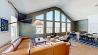 Located at 111 Cochran Rd in Clemson, South Carolina, this single-floor space offers a well-designed layout ideal for gathering and collaboration. The standout feature is the living room, which boasts soaring vaulted ceilings and a dramatic floor-to-ceiling window wall that floods the interior with natural light and frames views of the surrounding neighborhood. A large sectional leather sofa, wall-mounted television, and warm wood flooring give the area a comfortable yet polished feel. Two separate dining areas provide flexible options for group seating and meetings. One dining space features a round table with plaid upholstered chairs nestled beside bright corner windows, while the other functions as a more intimate meeting room equipped with a whiteboard, round table, and additional natural light from a high window. A connecting hallway links the spaces seamlessly, featuring an open seating nook with midcentury-style lounge chairs, exposed ductwork as an industrial design accent, and interior windows that offer views between rooms. Warm-toned hardwood flooring runs consistently throughout, tying the spaces together. The overall ambiance is modern, welcoming, and versatile, balancing communal functionality with thoughtfully curated furnishings and abundant natural light.