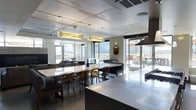 This single-floor space showcases a thoughtfully designed open-plan layout that seamlessly integrates the kitchen, dining, and living areas into one cohesive environment. The exposed concrete ceiling lends an industrial character to the space, complemented by statement pendant light fixtures featuring warm amber-toned panels that add a decorative focal point overhead.

The kitchen is equipped with built-in appliances and flows effortlessly into a generous dining area anchored by a long marble-topped table surrounded by amber-tinted chairs. A dark, substantial island serves as a transitional element between the kitchen and living spaces, providing additional surface area and casual seating.

The living area features a large sectional sofa in charcoal gray, accented with mustard yellow cushions, positioned around a bold teal feature wall displaying elegant artwork. Warm wood paneling frames the area, adding depth and texture to the interior palette.

Large windows and glass sliding doors flood the space with natural light while offering views of an outdoor terrace and the surrounding cityscape beyond, creating a strong visual connection between the interior and exterior environments.